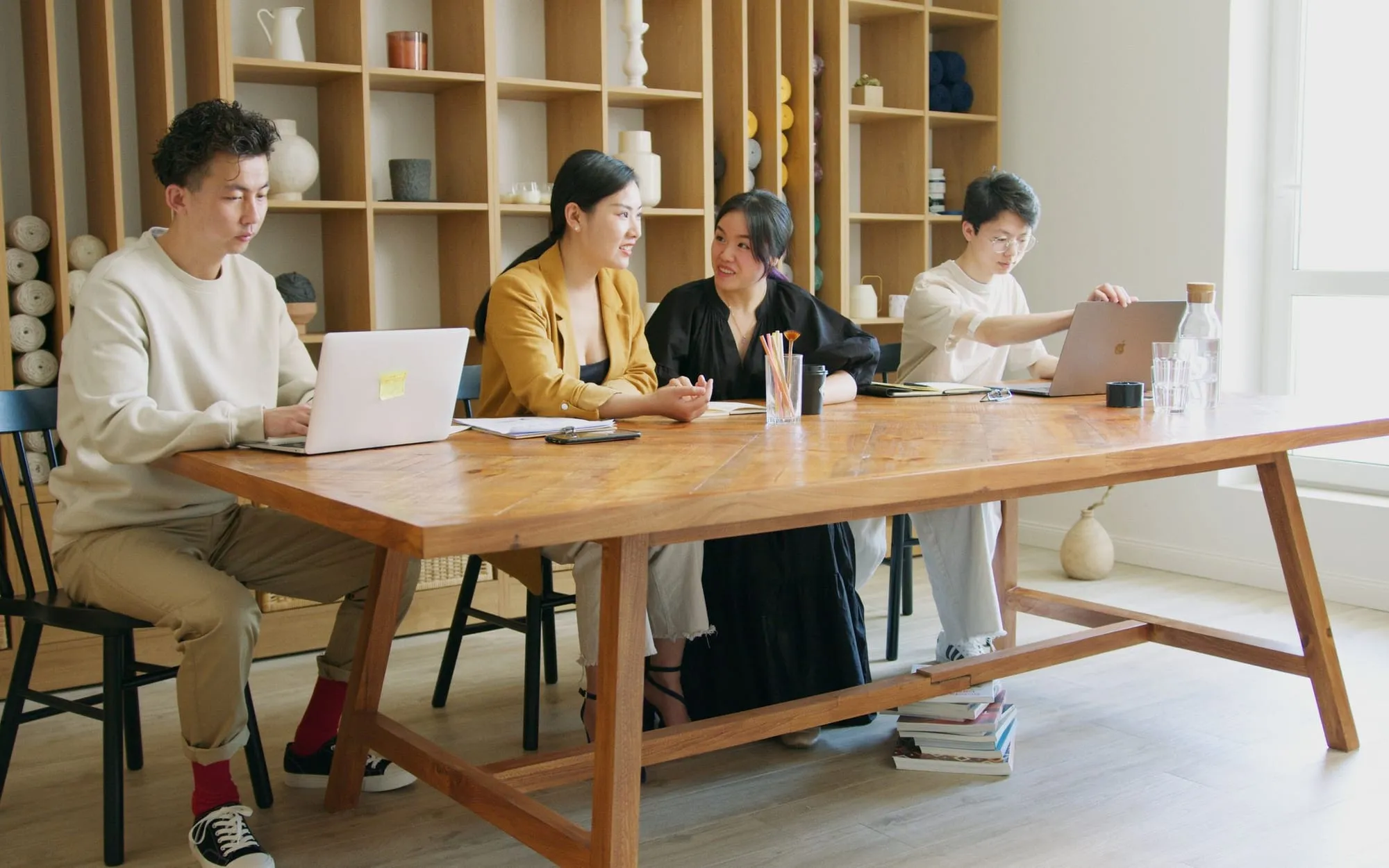 A team working on laptops around a long wooden table in a bright room lined with shelves