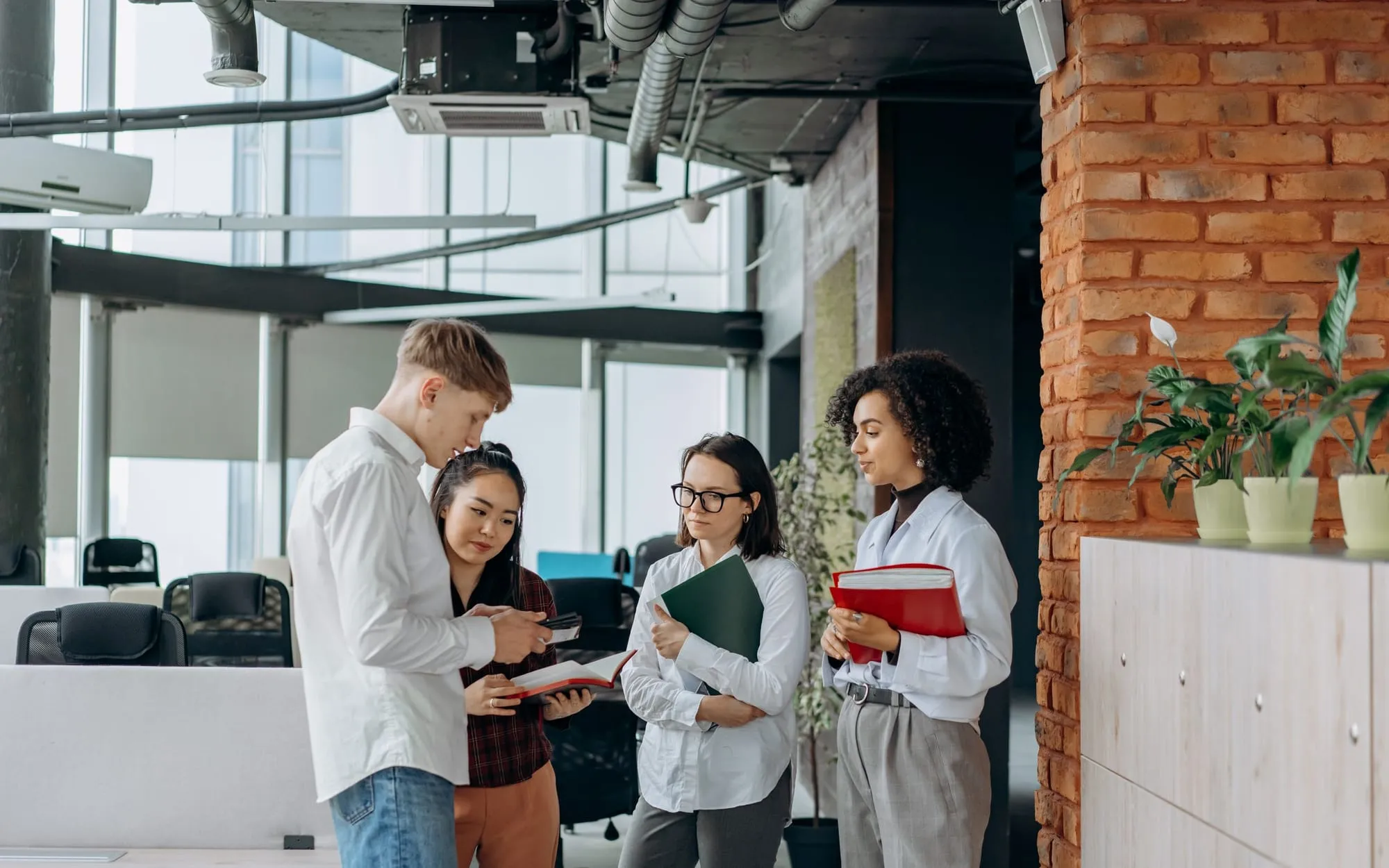 Four colleagues in white shirts reviewing notebooks and folders together in a modern office with a brick column