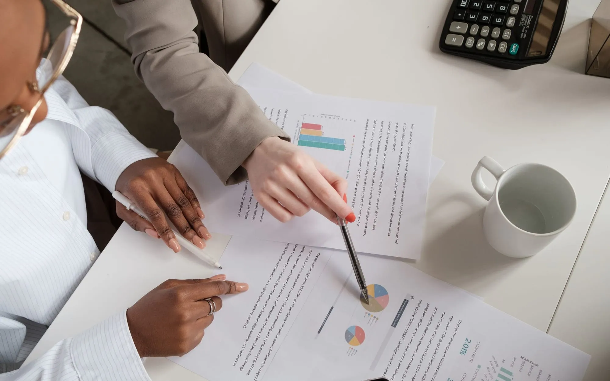 Hands pointing at printed charts and reports on a white desk, beside a mug and a calculator