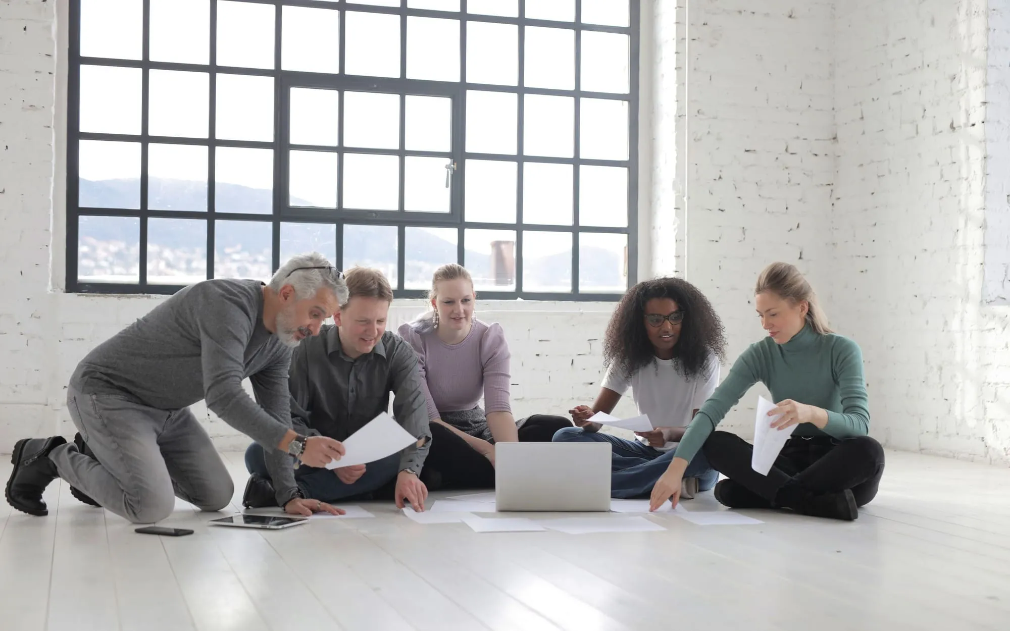 Colleagues kneeling and sitting on the floor of a bright loft office with tall windows, sharing papers and a laptop