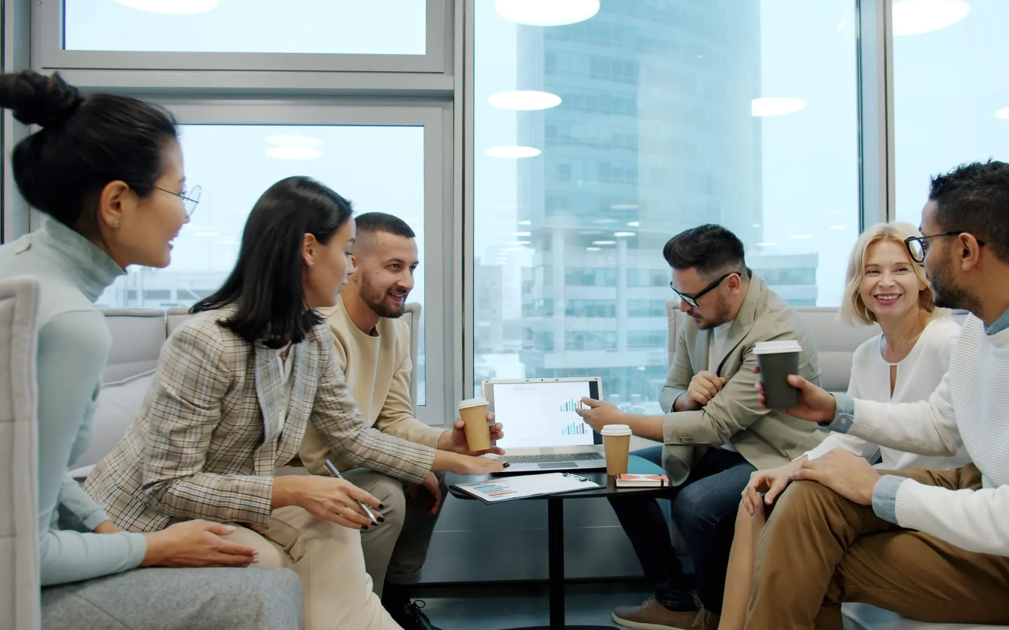 Six colleagues with coffee cups gathered around a laptop by tall windows in a high-rise office