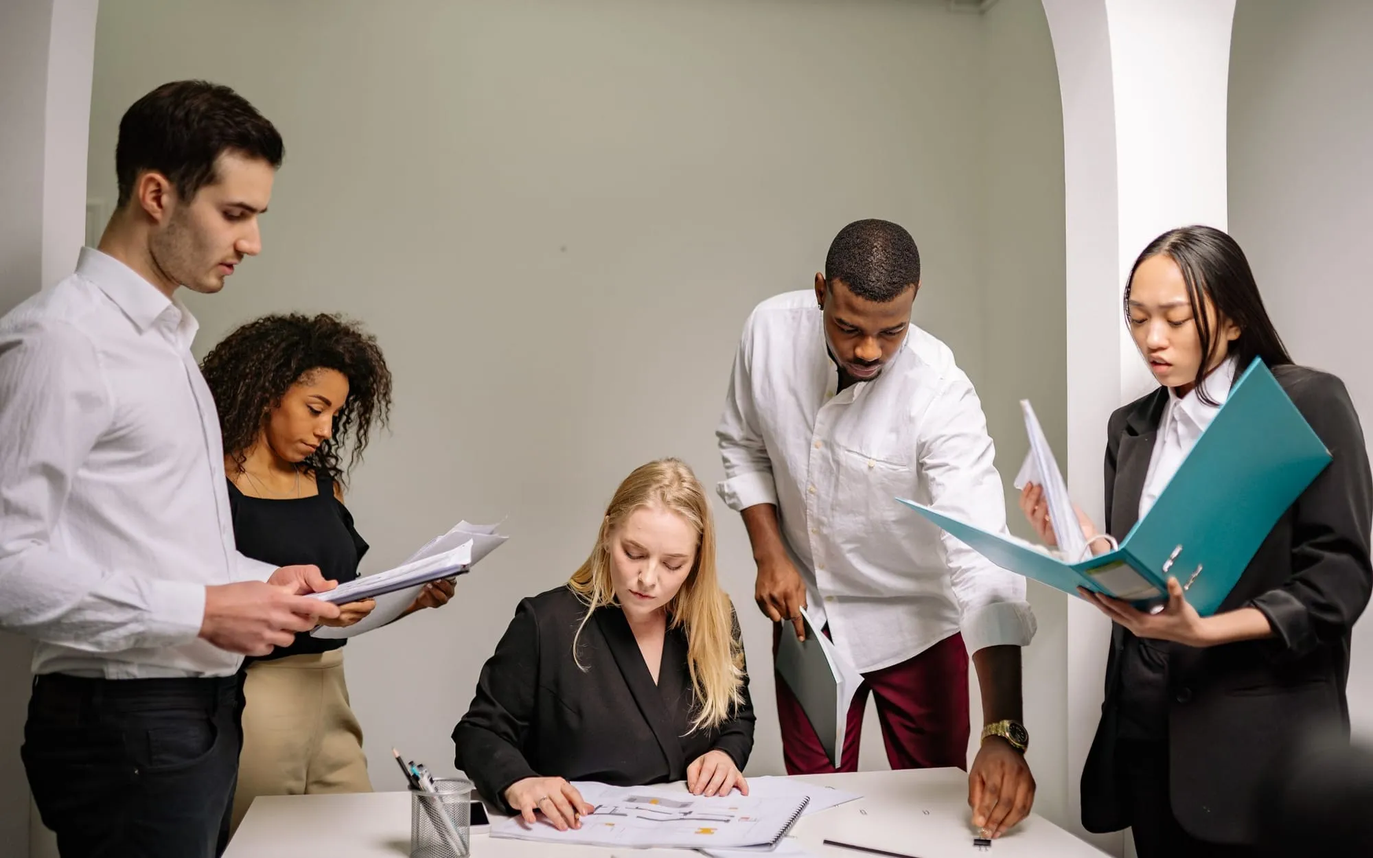 A team of five reviewing documents and a teal binder around a desk in a white office