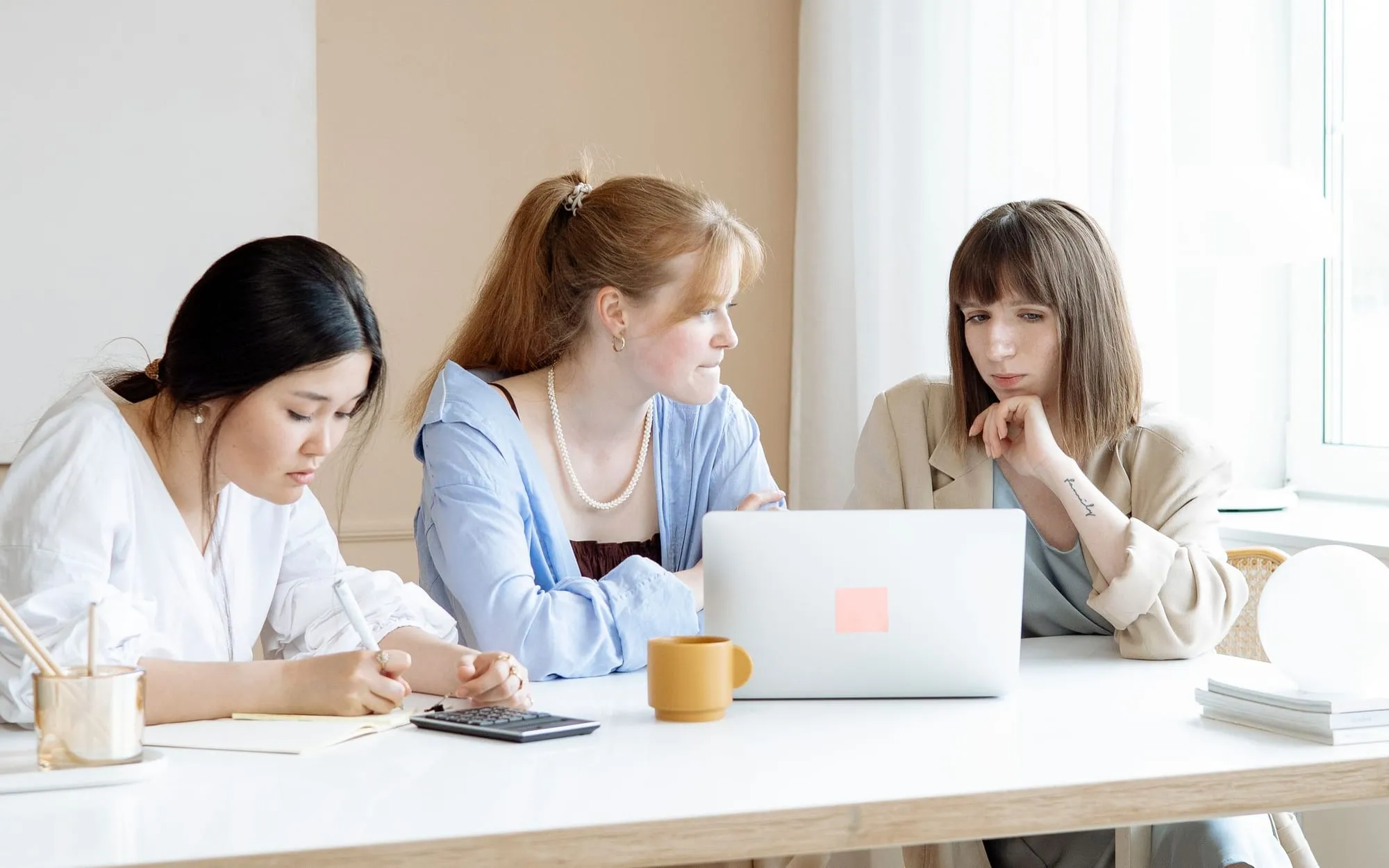 Three women at a white table looking over their notes and a shared laptop