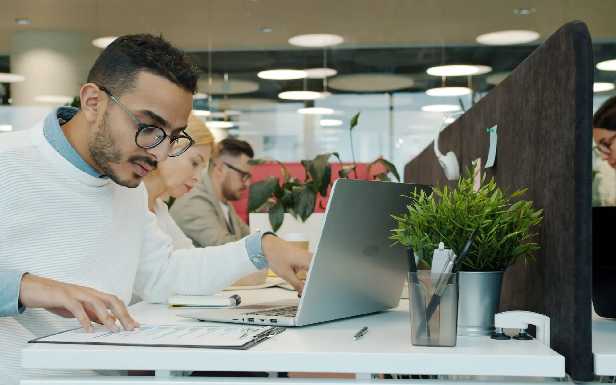 A man in glasses typing on a laptop at a row of desks in a busy open-plan office