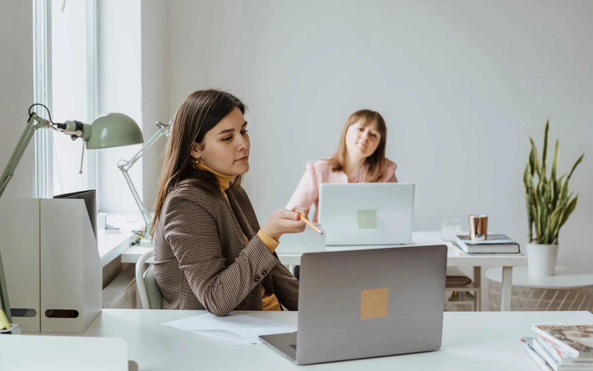 Two women working on laptops at white desks in a bright office, one pointing a pencil at her screen