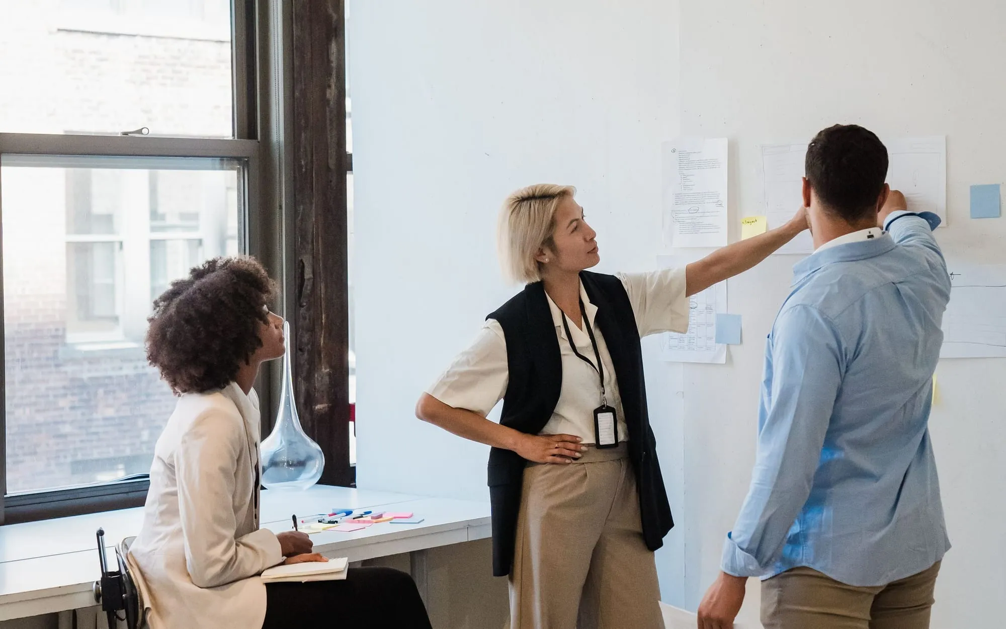 Three colleagues planning at a wall of pinned notes in a bright office, one of them seated with a notebook